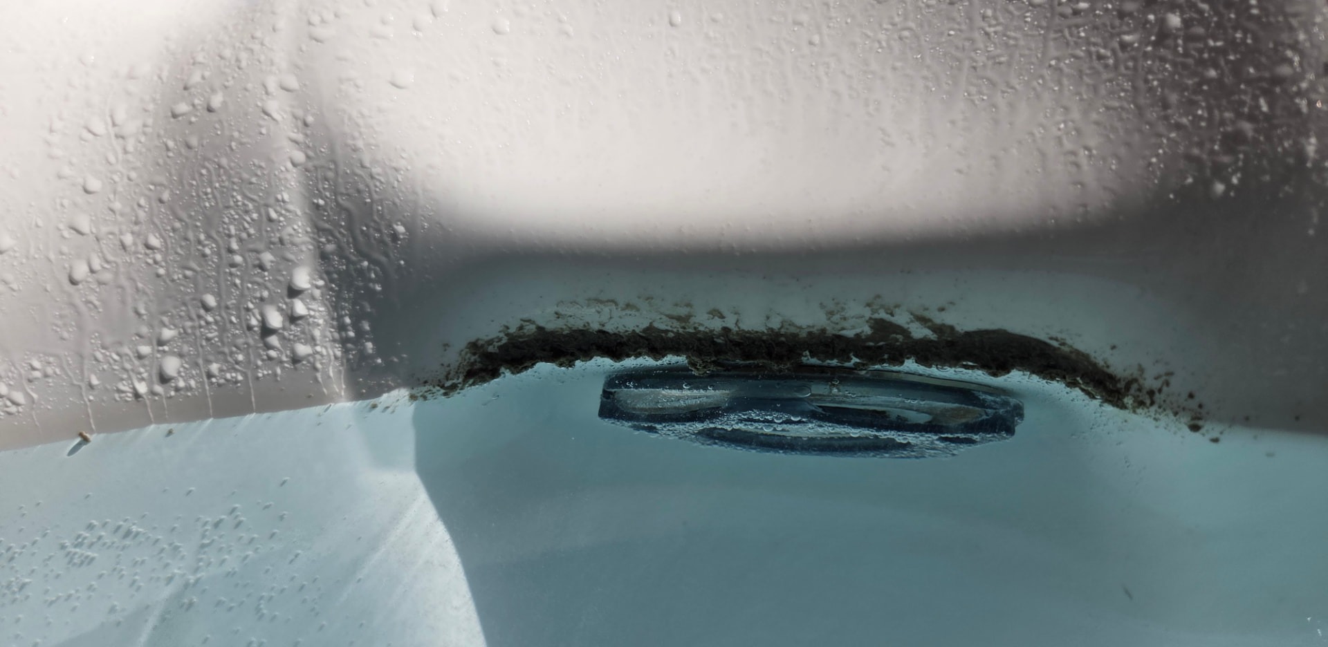 Biofilm buildup along a hot tub waterline: dark residue clinging to the underside of the white shell ledge above the water, next to a submerged jet fitting